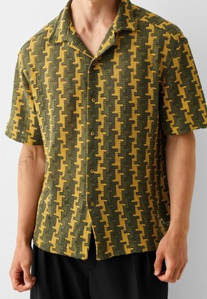 Mann trägt ein olivgrünes, gestricktes kurzärmeliges Hemd mit gelbem geometrischem Muster und schwarze Hose, steht vor einem einfarbigen Hintergrund.