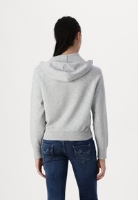 Femme aux cheveux tressés portant un sweat à capuche gris clair et un jean bleu foncé, vue de dos sur un fond blanc uni.