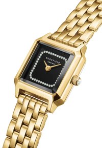 Reloj metálico dorado con una esfera negra cuadrada, bordeada de cristales transparentes. Presenta una correa texturizada y una corona con un pequeño acento de gema.