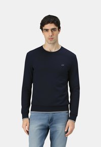 Marineblaues Sweatshirt aus glattem Stoff, mit Rundhalsausschnitt, gerippten Bündchen und einem dezenten Logo auf der Brust. Kombiniert mit hellblauen Jeans.