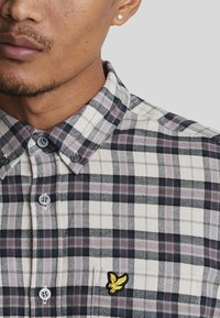 Geruite shirt in beige, paars en marineblauw, met een button-up kraag en geborduurd goudkleurig logo op de borstzak; de stof lijkt zacht en textuur te hebben.