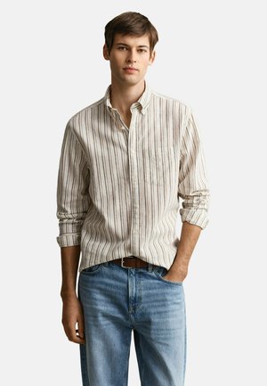 Junger Mann trägt beige-weiß gestreiftes Hemd mit hochgekrempelten Ärmeln, in hellblaue Jeans mit braunem Gürtel gesteckt, steht entspannt.