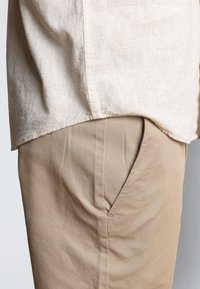 Homme portant un pantalon beige avec une main dans la poche et une chemise ample en lin blanc cassé non rentrée sur la taille.