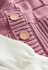 Gros plan d’un vêtement tricoté rose avec des motifs texturés et deux boutons ronds aux bords dorés tressés à côté d’un tissu blanc.