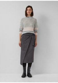 Frau trägt einen weichen grau-rosa Pullover, dunkelgrauen Wickelrock, schwarze Strumpfhosen und schwarze Stiefeletten, steht vor einer schlichten grauen Wand.