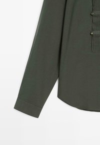 Chemise à manches longues vert foncé avec un panneau avant plissé et deux boutons métalliques sur fond blanc.