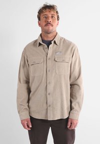 Camisa de pana beige con cierre de botones en la parte delantera, bolsillos en el pecho y un parche con el logo. Cuenta con un material estriado texturizado y un corte relajado.
