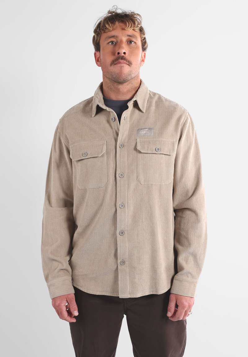Camisa de pana beige con cierre de botones en la parte delantera, bolsillos en el pecho y un parche con el logo. Cuenta con un material estriado texturizado y un corte relajado.