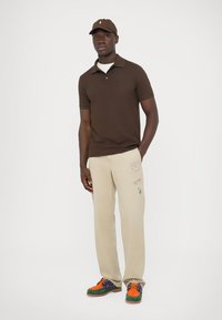 Barna texturált galléros póló, bézs színű nadrág hímzett részletekkel, és színes sportcipők. Tartozik hozzá egy hozzáillő barna sapka.