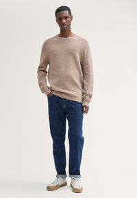 Beige stickad tröja med rund halsringning, kombinerad med mörkblå denimjeans. Vita sneakers med beige sulor, stående mot en neutral bakgrund.