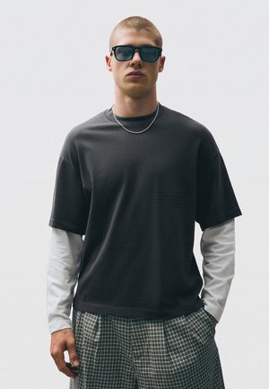 Jeune homme aux cheveux courts portant des lunettes de soleil foncées, un T-shirt noir superposé à une chemise blanche à manches longues, une chaîne argentée et un pantalon à carreaux.