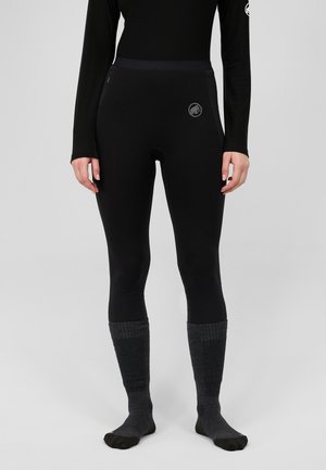 Person steht, trägt schwarze enganliegende Leggings und langärmliges Oberteil mit dunkelgrauen knielangen Socken vor einem schlichten hellen Hintergrund.