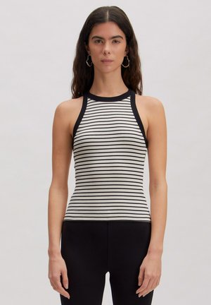 Vrouw met lang donker haar die oorringen draagt, een mouwloos wit en zwart gestreept topje en zwarte broek, staand tegen een effen achtergrond.