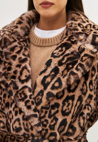 Abrigo de piel sintética de estampado de leopardo con un amplio cuello, textura suave y cintura ceñida con cinturón, llevado sobre un suéter de punto beige.