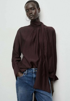 Mujer con una blusa de manga larga marrón oscuro con un detalle de bufanda, combinada con jeans de mezclilla azules, de pie contra una pared clara.
