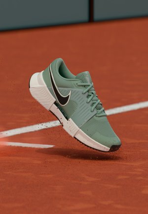 Pantof sport verde Nike cu logo negru în formă de swoosh plutind deasupra terenului de tenis cu zgură roșie, praf roșu de zgură împrăștiindu-se lângă linia albă a terenului.