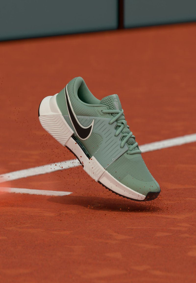 Baskets Nike vertes avec le logo noir en mouvement au-dessus d'un court de tennis en terre battue, de la poussière rouge se dispersant près de la ligne blanche du court.