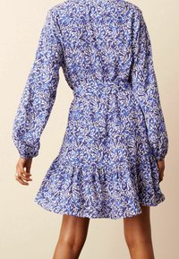 Abito a fiori blu e bianchi con maniche lunghe, vita cintata e orlo arricciato, realizzato in tessuto leggero.