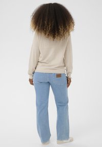 Ženska s kodrasto pričesko, oblečena v bež pulover, svetlo modre kavbojke z zadnjimi žepi in bež čevlje, stoji pred belo ozadjem.