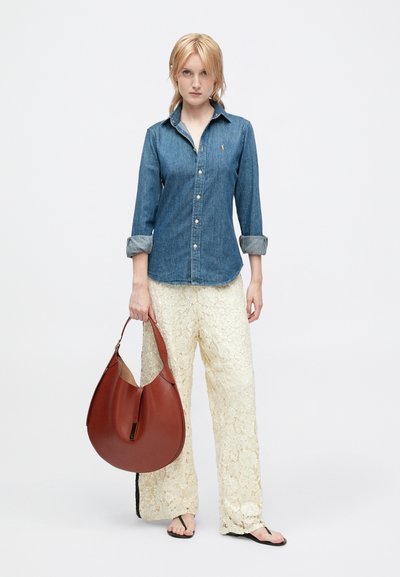 Ženska nosi jeans srajco, široke hlače iz kremaste čipke, črne sandale in drži veliko rjavo usnjeno torbico za ročaj.