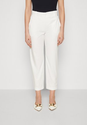 Vrouw die witte, hoogverstige cropped broek draagt, gecombineerd met witte schoenen met puntige neus en gouden kettinkdetail.