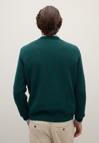 Grüner Pullover mit strukturierter Oberfläche, geripptem Kragen und Bündchen, von hinten gezeigt. Kombiniert mit hellen Hosen, die ein schlichtes Design präsentieren.