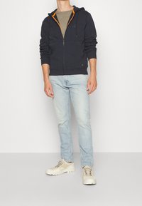 Sudadera con cremallera en color navy, con detalles en naranja, llevada sobre una camiseta gris. Combinada con jeans de color azul claro y zapatos beige. Textura suave, ajuste casual.