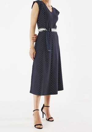 Femme portant une robe midi bleu marine avec des rayures diagonales blanches, une ceinture blanche avec boucle noire et des sandales noires à talons hauts.