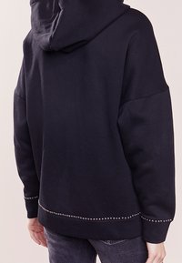 Sudadera negra con una textura suave, que presenta una capucha y detalles de tachuelas a lo largo del dobladillo inferior. Diseño holgado con hombros caídos.