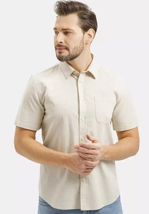 Mężczyzna z brodą, ubrany w beżową, krótką koszulę na guziki, złożone ręce, patrzący w lewo na jednolitym tle.