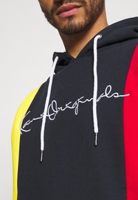 Mikina s kapucí v námořnické, červené a žluté barevné bloky, s bílým vyšitým logem a bílými šňůrkami. Měkký, přes hlavu.