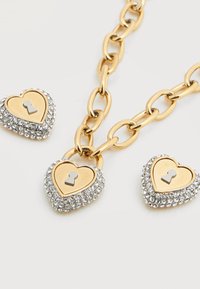 Κολιέ με χρυσή αλυσίδα που διαθέτει τρία μενταγιόν σε σχήμα καρδιάς, το καθένα με χρυσό κέντρο και περιβαλλόμενο από ασημένιους κρυστάλλους, με έντονη υφή και λάμψη.