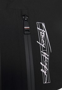 Černá zapínací bunda s bílým podpisovým logem. Tkanina je hladká s matným povrchem a poblíž zipu je barevný pruh jako akcent.