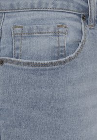 Un jean en denim bleu clair présente une poche arrière avec des coutures visibles et des détails en rivets, mettant en avant une texture lisse et un design classique.