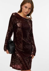 Burgunderfarget paljettkjole med lange ermer og rund hals, med en tettsittende silhuett. Modellen holder en svart clutchveske.
