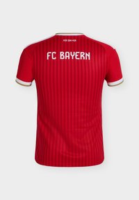 Κόκκινη φανέλα ποδοσφαίρου της FC Bayern με κατακόρυφες ρίγες, λευκές και χρυσές λεπτομέρειες στα μανίκια και το κείμενο "FC BAYERN" και "MIA SAN MIA" στην πλάτη.