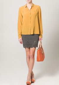 Blusa gialla con bottoni e maniche lunghe, abbinata a una gonna grigia con bordi scallop e dettagli a intaglio. Gli accessori includono una borsa arancione.