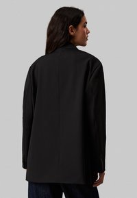 Blazer nero su misura con silhouette strutturata, texture liscia, cucitura posteriore sottile e dettagli con bottoni sui polsini delle maniche.