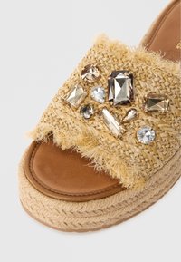 Geflochtene Espadrille-Sandale in natürlichem Tan mit Fransenakzenten, ausgestattet mit einer Anordnung verschiedener klarer und brauner Edelsteine in unterschiedlichen Größen auf dem Riemen.