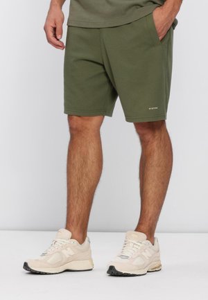 Uomo che indossa pantaloncini color verde oliva con trama e sneakers beige New Balance, in piedi con una mano in tasca su uno sfondo semplice.