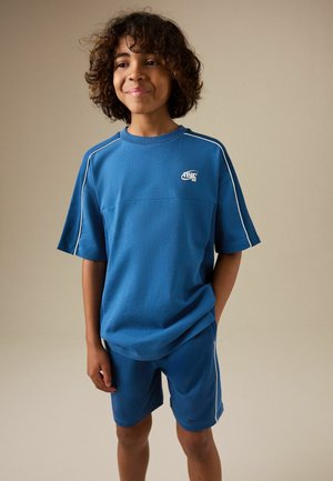 Garçon aux cheveux bouclés portant une chemise bleue à manches courtes et un short assorti avec un liseré blanc, debout devant un fond neutre.