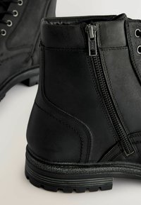 Las botas de cuero negro a nivel del tobillo presentan un cierre lateral con cremallera y un diseño de cordones. La suela es texturizada, lo que proporciona agarre, con detalles de costura visibles.