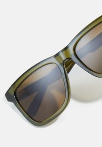Ochelari de soare supradimensionați, cu nuanță verde, ram�ă transparentă și lentile degradate maro. Dispun de un design ușor din plastic și o formă unghiulară.