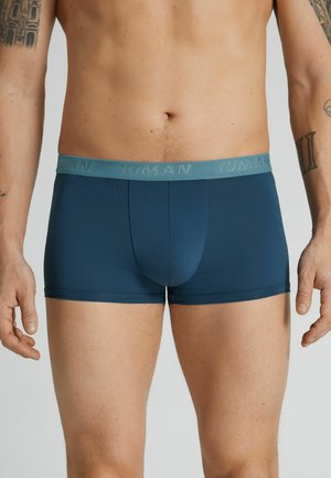 Mandekrop iført mørkeblå boksershorts med et turkis midjebånd med teksten "IUMAN", tatoverede arme synlige på siderne.