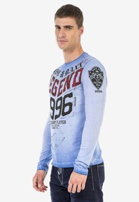 Langarmshirt aus einem gradienten blauen Stoff, mit auffälligem schwarzen und roten grafischen Text sowie einem Patch auf dem linken Ärmel. Weicher Stoff.