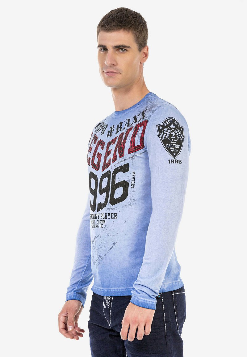 Langarmshirt aus einem gradienten blauen Stoff, mit auffälligem schwarzen und roten grafischen Text sowie einem Patch auf dem linken Ärmel. Weicher Stoff.