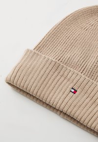 Beige strikket lue med ribbestruktur og brettet kant. Inkluderer et lite logo-patch i rød, hvit og blå på kanten.