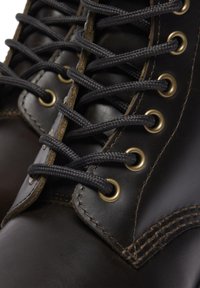 Botas de cuero marrón con cordones negros, ojales dorados y costuras visibles. El material es suave con un diseño resistente y puntera redondeada.