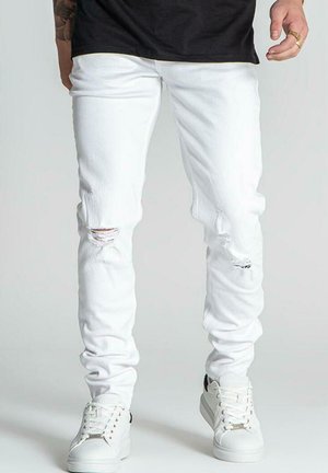 White denim skinny jeans with distressed rips at knees, textured fabric, and a slim fit, paired with white sneakers.
