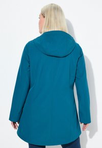 Kvinna med blont hår som bär en teal-färgad jacka med huva, vänd bort, stående mot en enkel vit bakgrund.